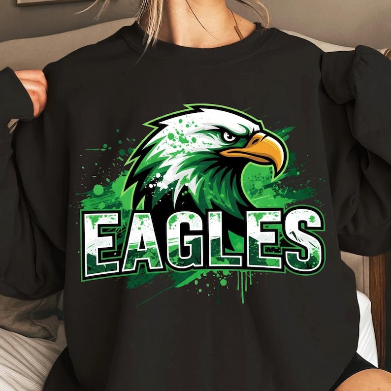 Op de afbeelding: Zwart sweatshirt met een groen en wit adelaarsontwerp en het woord "EAGLES". De afbeelding bevat groene verfspatten. De adelaar heeft een gele snavel en een geconcentreerde uitdrukking. Een sportief ontwerp.