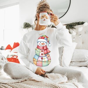 Puede incluir: Sudadera blanca con un dise&ntilde;o de mu&ntilde;eco de nieve colorido. El mu&ntilde;eco de nieve tiene un cuerpo de retazos, una bufanda roja y rosa y un sombrero festivo. El dise&ntilde;o incluye una guirnalda de luces. Se sostiene una taza de Pap&aacute; Noel frente a la cara.