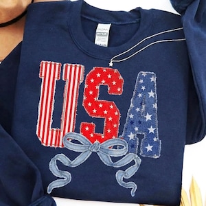 May include: Navy blue sweatshirt with the word "USA" appliqued in red, white, and blue fabric. The "U" features red and white stripes, the "S" has red stars, and the "A" has blue stars. A light blue bow is sewn below the letters.