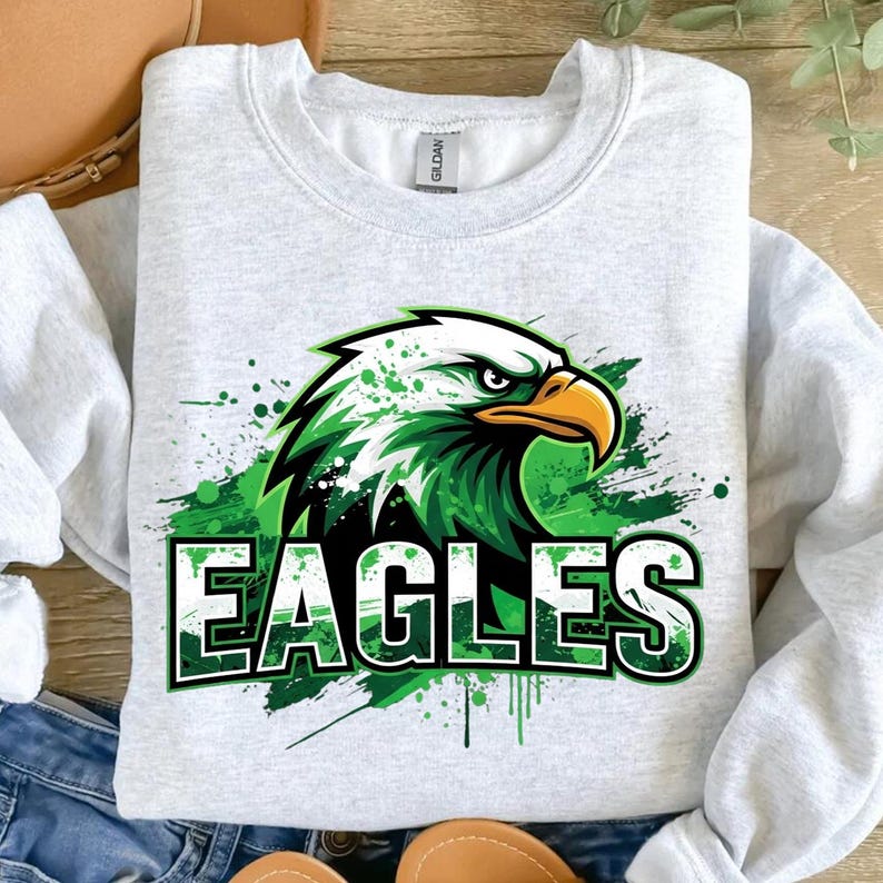 Op de afbeelding: Een lichtgrijs sweatshirt met een groen en witte adelaar en het woord "EAGLES" in het groen. De adelaarsafbeelding heeft een spatteffect. De sweatshirt is gemaakt van een zacht materiaal.