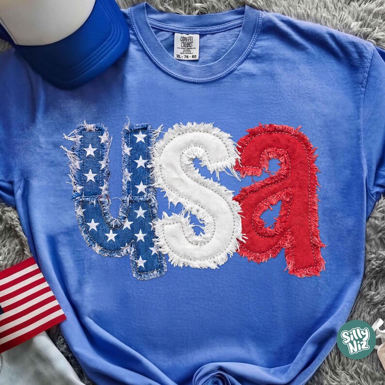 May include: A blue t-shirt with the word "USA" appliqued on the front. The letters are in red, white, and blue, with the "U" and "S" in denim with white stars. The "A" is red. A blue baseball cap is visible in the background.