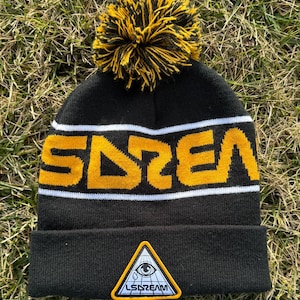 Peut inclure: Un bonnet noir avec un pompon jaune et noir sur le dessus. Le bonnet a une bande blanche avec le mot "DREAM" en lettres jaunes. Le bonnet a également un patch noir et blanc avec un triangle et un œil à l'intérieur, et les mots "LSDREAM" sous l'œil.