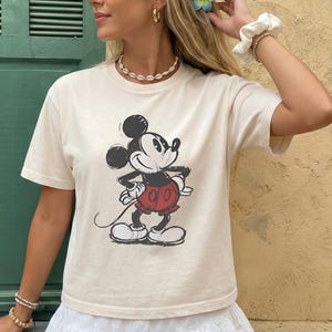 Puede incluir: Camiseta de color marfil con un gr&aacute;fico cl&aacute;sico de Mickey Mouse en negro, rojo y blanco. La camiseta tiene cuello redondo y mangas cortas. La palabra "IVORY" est&aacute; impresa en la esquina inferior izquierda de la imagen.