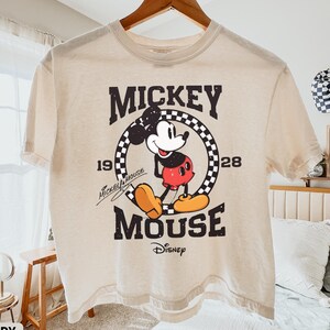 May include: Ivory-colored t-shirt featuring a vintage Mickey Mouse graphic. The design includes the words "Mickey Mouse" and "Disney" along with the years "19" and "28". The shirt is hanging on a wooden hanger.