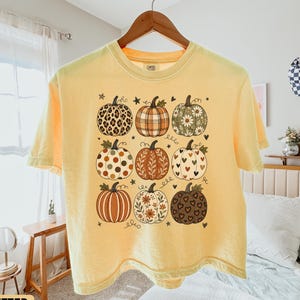 May include: A pale yellow short-sleeve t-shirt featuring a graphic print of nine pumpkins arranged in a grid. Each pumpkin displays a unique pattern, including leopard print, plaid, floral motifs, polka dots, and heart shapes, accented by small stars.