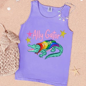 Può includere: Una canotta viola con la scritta "Ally Gator Pride" e una grafica di un alligatore verde con un mantello arcobaleno. La canotta è su una superficie sabbiosa con una borsa intrecciata e una collana di conchiglie.