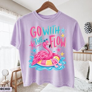 May include: A light purple t-shirt featuring a graphic of a pink flamingo floating on a yellow and pink inner tube in blue water. The text "GO WITH THE FLOW" is printed above the flamingo in bright pink and blue letters, surrounded by small pink and blue stars.