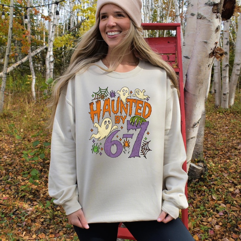 May include: A beige sweatshirt with the text "HAUNTED BY 6-7" in orange, green, and purple. The design includes ghosts, spiderwebs, and a spider. The sweatshirt is a casual, long-sleeved garment.