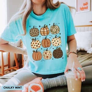 May include: A bright turquoise t-shirt featuring a graphic print of nine pumpkins in a three-by-three grid. Each pumpkin displays a unique pattern, including leopard print, plaid, floral motifs, polka dots, and heart shapes, accented by small stars.