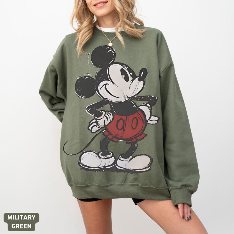 Puede incluir: Sudadera verde militar con un gr&aacute;fico desgastado de Mickey Mouse en negro, blanco y rojo. La sudadera tiene mangas largas y cuello redondo. El texto "MILITARY GREEN" est&aacute; en la parte inferior izquierda.