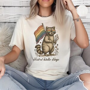 Op de afbeelding: Crèmekleurig T-shirt met een wasbeer met een zonnebril en een regenboogvlag. De wasbeer is omringd door bloemen en de tekst "Foxtrot Delta Tango" staat eronder. Het T-shirt wordt door een persoon gedragen.