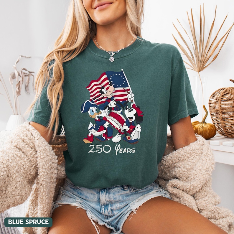 May include: Blue spruce t-shirt featuring a graphic of cartoon characters holding a flag with red and white stripes and a blue field with white stars. The text "250 Years" is below the graphic. The shirt is a relaxed fit.