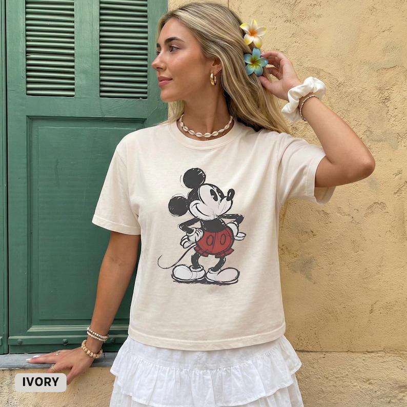 Puede incluir: Camiseta de color marfil con un gr&aacute;fico cl&aacute;sico de Mickey Mouse en negro, rojo y blanco. La camiseta tiene cuello redondo y mangas cortas. La palabra "IVORY" est&aacute; impresa en la esquina inferior izquierda de la imagen.