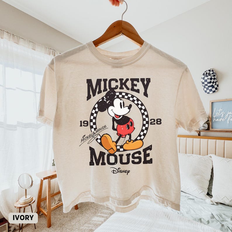 May include: Ivory-colored t-shirt featuring a vintage Mickey Mouse graphic. The design includes the words "Mickey Mouse" and "Disney" along with the years "19" and "28". The shirt is hanging on a wooden hanger.
