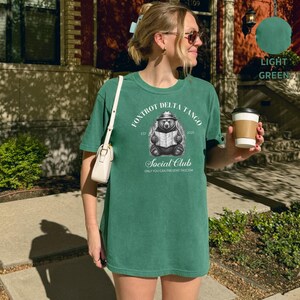 Puede incluir: Una camiseta verde con un gr&aacute;fico de un oso con un sombrero y sosteniendo un mapa. El texto en la camiseta dice "Fox Trot Delta Tango Est 2025 Social Club Only You Can Prevent Fascism".