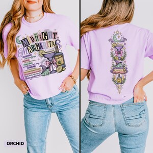 May include: A light purple t-shirt featuring a graphic print with the text Midnight Margaritas, a margarita glass, a tequila bottle, and a frog wearing a witch hat. The back of the shirt displays a vertical design with a crescent moon, a margarita glass, a frog on books, and a tequila bottle surrounded by stars and foliage.