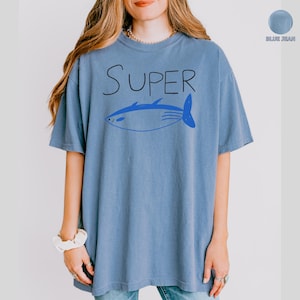 May include: Blue jean coloured t-shirt featuring the word "SUPER" in black script above a blue fish illustration. The shirt is short-sleeved and casual.