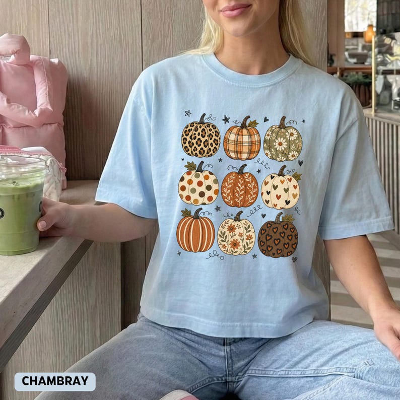 May include: A light blue short-sleeve t-shirt featuring a graphic print of nine pumpkins arranged in a grid. Each pumpkin displays a unique pattern, including leopard print, plaid, polka dots, floral motifs, and heart shapes in shades of orange and brown.