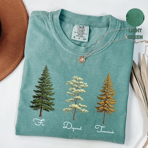 Può includere: Una maglietta verde chiaro con un lavaggio vintage che presenta tre alberi con i nomi "Fir", "Dogwood" e "Tamarack" stampati sotto. La maglietta ha un'etichetta che recita "Comfort Colors" e "S - P - CH".