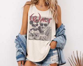 May include: An ivory sleeveless tank top featuring a graphic print of a man and a cartoon mouse both wearing sunglasses. The text "WALT" is printed in a large, stylized maroon font above the figures. The garment has a scoop neckline and wide straps.