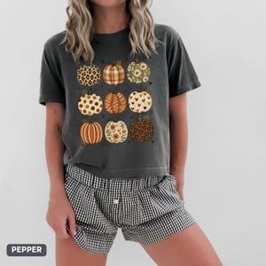 May include: A charcoal gray short-sleeve t-shirt featuring a graphic print of nine pumpkins in a grid layout. Each pumpkin displays a unique pattern, including leopard print, plaid, floral motifs, polka dots, and heart shapes in earthy tones.