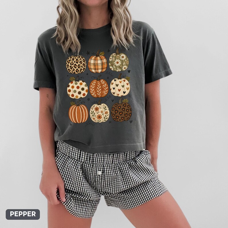 May include: A charcoal gray short-sleeve t-shirt featuring a graphic print of nine pumpkins in a grid layout. Each pumpkin displays a unique pattern, including leopard print, plaid, floral motifs, polka dots, and heart shapes in earthy tones.