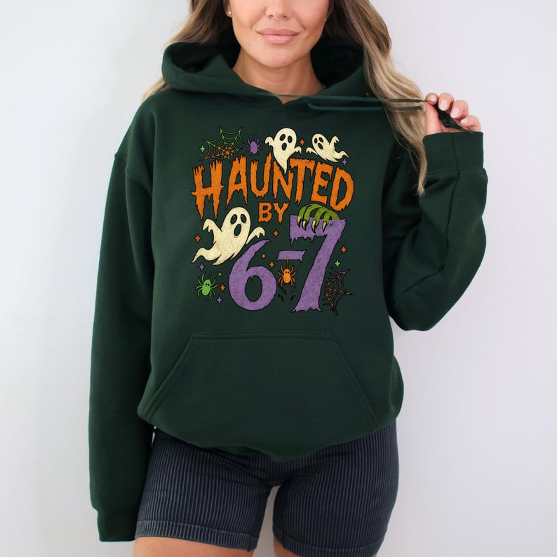 May include: Dark green hooded sweatshirt with a Halloween-themed graphic. The design features the text "HAUNTED BY 6-7" in orange and purple, along with ghost and spiderweb illustrations. The sweatshirt has a front pocket and a drawstring hood.