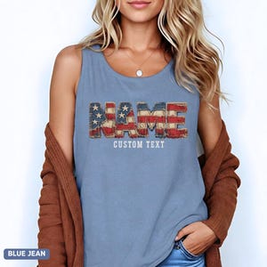 Puede incluir: Una camiseta sin mangas azul con la palabra NAME en un patrón de bandera estadounidense desgastada con estrellas y rayas. Debajo de la palabra, el texto CUSTOM TEXT está impreso en blanco. La prenda se lleva con un cárdigan marrón y vaqueros.