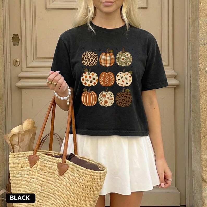 May include: A black short-sleeve t-shirt featuring a graphic print of nine pumpkins arranged in a grid. Each pumpkin displays a unique pattern, including leopard print, plaid, floral, polka dots, leaves, hearts, and solid orange stripes.