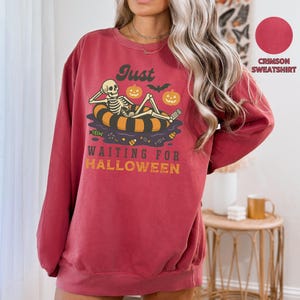 May include: Crimson sweatshirt with a Halloween-themed graphic. The design features a skeleton relaxing on an inner tube with pumpkins and the text "Just waiting for Halloween". The sweatshirt is a comfortable, casual style.