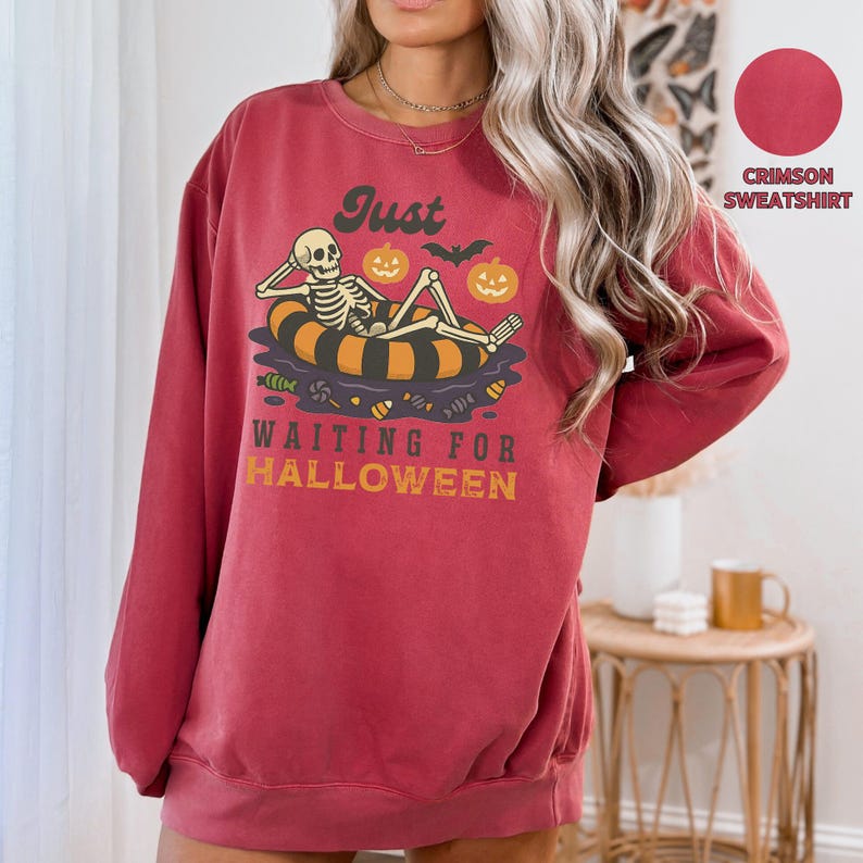 May include: Crimson sweatshirt with a Halloween-themed graphic. The design features a skeleton relaxing on an inner tube with pumpkins and the text "Just waiting for Halloween". The sweatshirt is a comfortable, casual style.