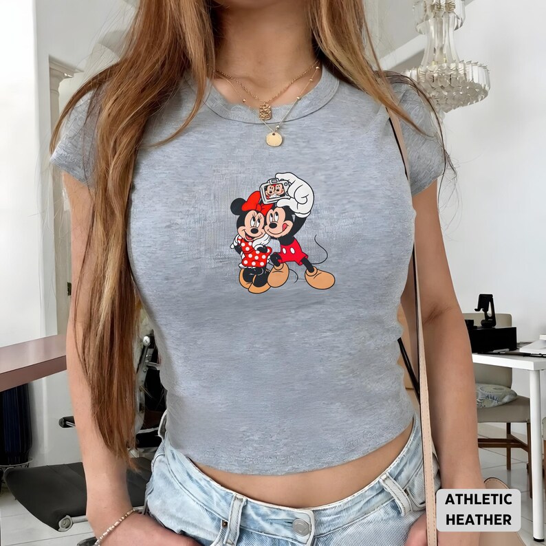 May include: Athletic heather gray crop top featuring a colorful graphic of Mickey and Minnie Mouse. Minnie wears a red polka dot dress, while Mickey holds a camera. The shirt has a crew neck and short sleeves.