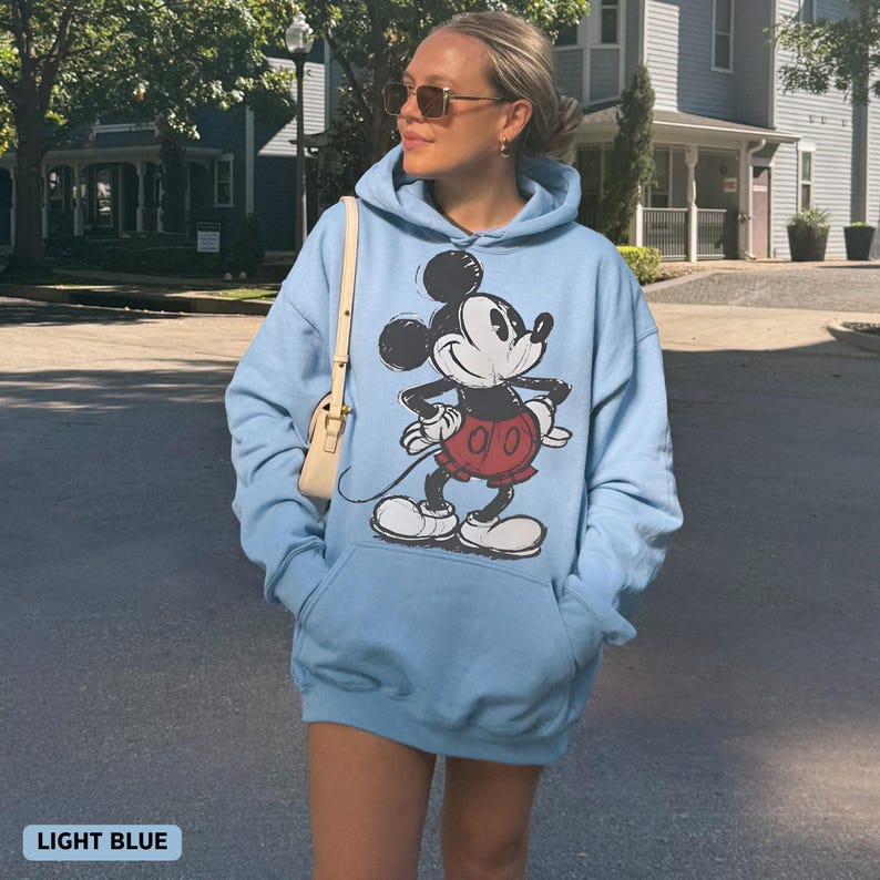Puede incluir: Sudadera con capucha azul claro con un dise&ntilde;o de Mickey Mouse. La sudadera tiene un bolsillo de canguro y una capucha con cord&oacute;n. El texto "LIGHT BLUE" est&aacute; en la parte inferior de la imagen.