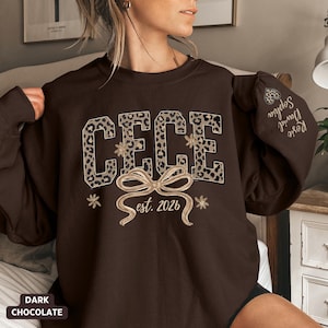 May include: Dark chocolate-colored sweatshirt with the name "CECE" in leopard print letters. Below the name is a bow and the text "est. 2026". The sleeve has the names "Rosie, David, Sophia" embroidered on it. The words "Dark Chocolate" are at the bottom.