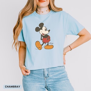 Puede incluir: Camiseta azul claro con un gr&aacute;fico de Mickey Mouse. El personaje de dibujos animados est&aacute; representado con pantalones cortos rojos, zapatos amarillos y orejas negras. El logotipo de Disney est&aacute; impreso debajo. La palabra "CHAMBRAY" est&aacute; impresa en la parte inferior.