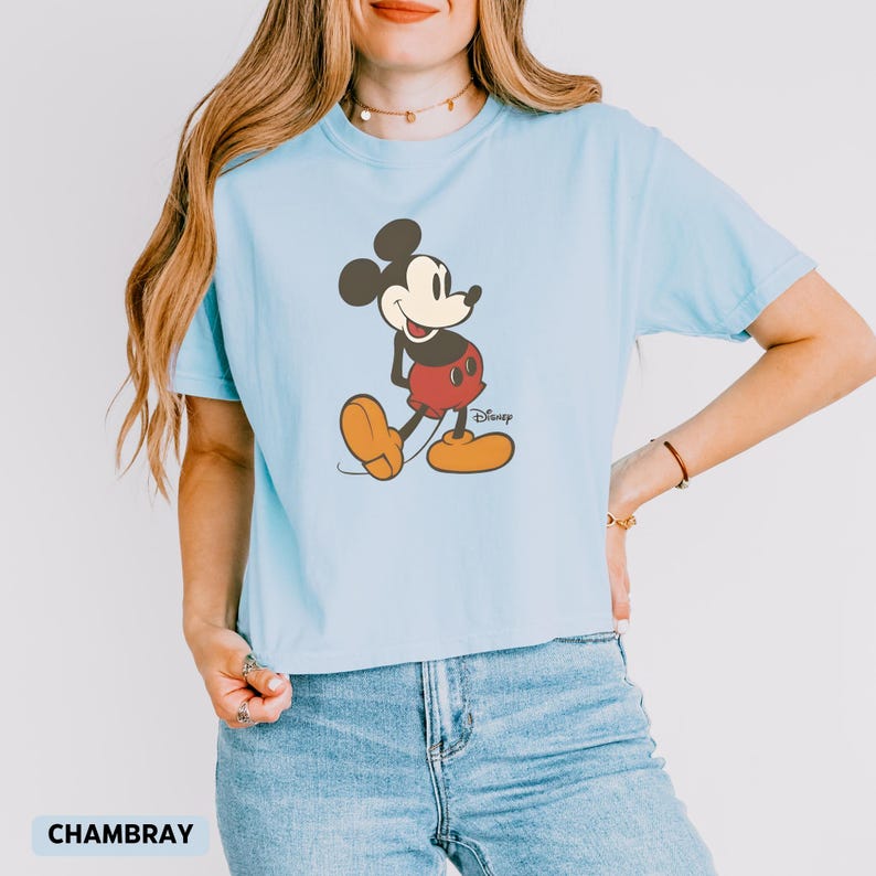 Puede incluir: Camiseta azul claro con un gr&aacute;fico de Mickey Mouse. El personaje de dibujos animados est&aacute; representado con pantalones cortos rojos, zapatos amarillos y orejas negras. El logotipo de Disney est&aacute; impreso debajo. La palabra "CHAMBRAY" est&aacute; impresa en la parte inferior.