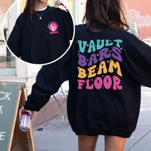 May include: Black sweatshirt with a graphic on the back that says "VAULT BARS BEAM FLOOR" in colorful, retro-style lettering. The front of the sweatshirt has a smaller graphic of a gymnast in a circle with the text "I AM AMAZING" and "SOPHIA".