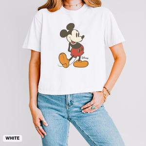 Puede incluir: Camiseta blanca con un dibujo de Mickey Mouse. El personaje de dibujos animados est&aacute; representado en negro, rojo y amarillo. El logotipo de Disney es visible. La camiseta es de estilo casual, de manga corta.