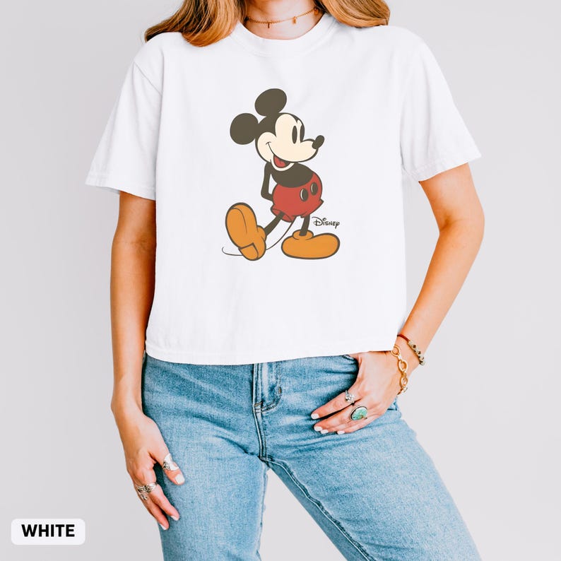 Puede incluir: Camiseta blanca con un dibujo de Mickey Mouse. El personaje de dibujos animados est&aacute; representado en negro, rojo y amarillo. El logotipo de Disney es visible. La camiseta es de estilo casual, de manga corta.