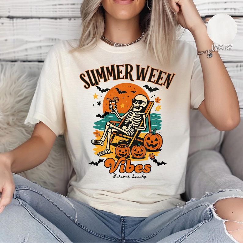 May include: Ivory-colored t-shirt with a graphic featuring a skeleton relaxing on a beach chair, holding a drink. The design includes pumpkins, bats, and the words "Summer Ween Vibes Forever Spooky."