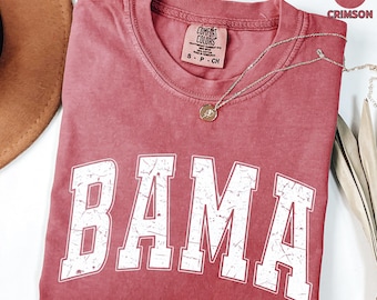 May include: Crimson-colored t-shirt with the word "BAMA" in distressed white lettering. The shirt has a crew neck and a small tag with text. A silver necklace with a pendant is draped across the shirt.
