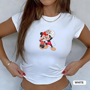 May include: White fitted t-shirt featuring a colorful graphic of Mickey and Minnie Mouse taking a selfie. Mickey is wearing red shorts and Minnie a red polka dot dress. The word "WHITE" is printed at the bottom.