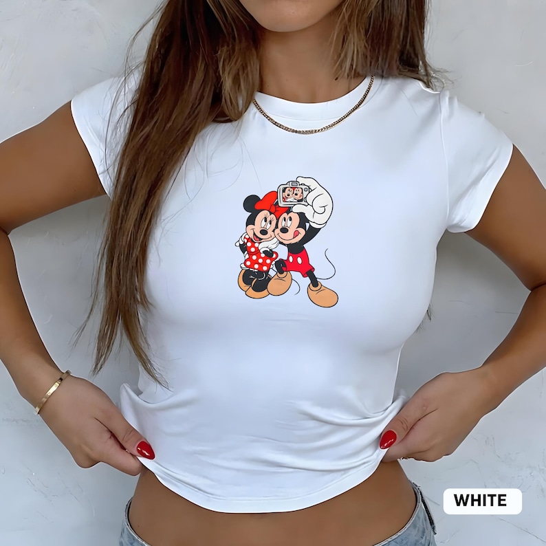 May include: White fitted t-shirt featuring a colorful graphic of Mickey and Minnie Mouse taking a selfie. Mickey is wearing red shorts and Minnie a red polka dot dress. The word "WHITE" is printed at the bottom.