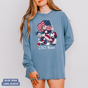 May include: Blue jean long-sleeve shirt featuring a graphic of Mickey Mouse, Donald Duck, and Minnie Mouse holding a flag with a red and white striped design and a blue field of stars. The text "250 Years" is below the characters.