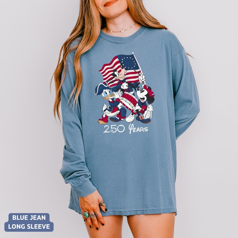 May include: Blue jean long-sleeve shirt featuring a graphic of Mickey Mouse, Donald Duck, and Minnie Mouse holding a flag with a red and white striped design and a blue field of stars. The text "250 Years" is below the characters.