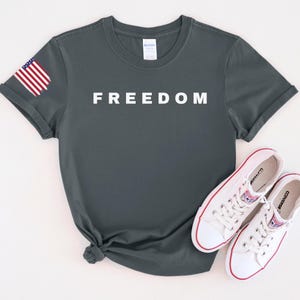 K&ouml;nnte beinhalten: Dunkelgraues T-Shirt mit dem Wort "FREEDOM" in wei&szlig;en Blockbuchstaben. Auf dem linken &Auml;rmel befindet sich eine kleine amerikanische Flagge. Wei&szlig;e Leinenschuhe mit roten Akzenten liegen darunter.