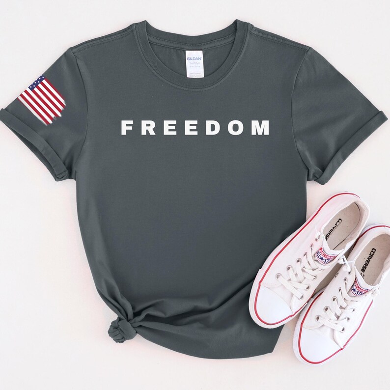 K&ouml;nnte beinhalten: Dunkelgraues T-Shirt mit dem Wort "FREEDOM" in wei&szlig;en Blockbuchstaben. Auf dem linken &Auml;rmel befindet sich eine kleine amerikanische Flagge. Wei&szlig;e Leinenschuhe mit roten Akzenten liegen darunter.