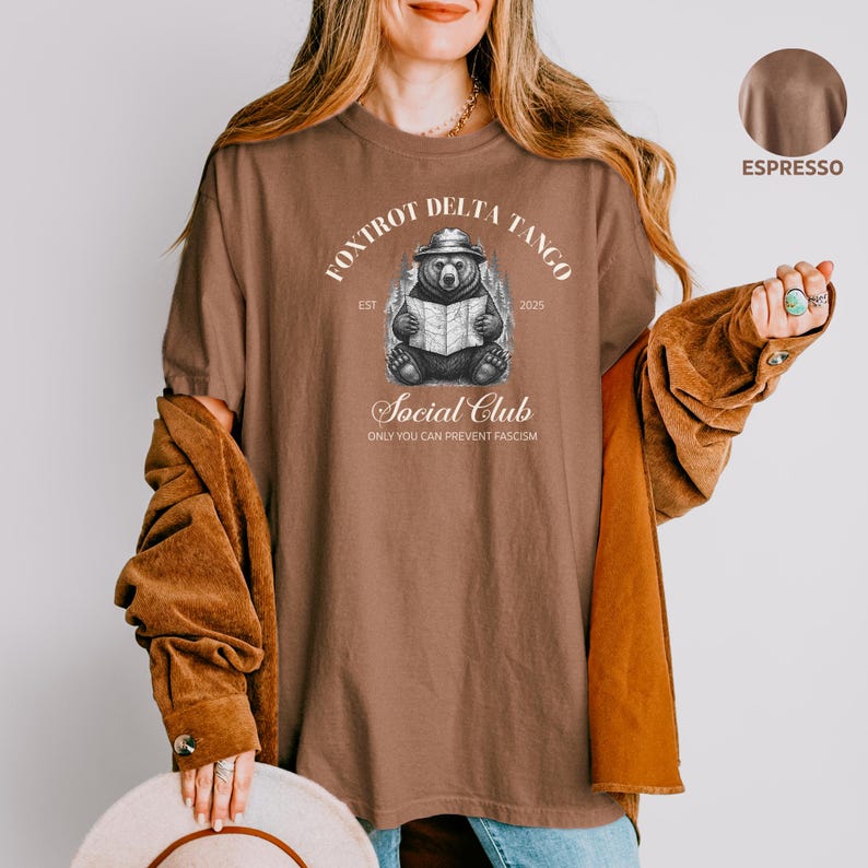Puede incluir: Una camiseta marr&oacute;n con un gr&aacute;fico de un oso con un sombrero leyendo un libro. El texto "Fox Trot Delta Tango Est 2025 Social Club Only You Can Prevent Fascism" est&aacute; impreso en la camiseta.