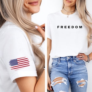 K&ouml;nnte beinhalten: Wei&szlig;es T-Shirt mit dem Wort "FREEDOM" in Schwarz aufgedruckt. Das Shirt hat auch ein kleines amerikanisches Flaggen-Design auf dem &Auml;rmel. Das Model tr&auml;gt blaue Jeans.