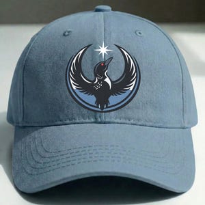 May include: A light blue baseball cap featuring a circular emblem with a black bird design, a white star, and a red eye. The bird is set against a blue background. The cap has a curved brim and an adjustable strap.